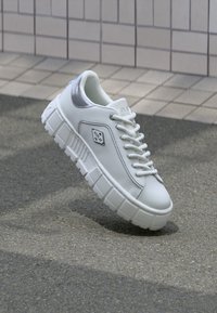 Valge nahast tennis, millel on paks ja tekstuuriga tall, hõbedased aktsendid ja rombikujuline logo näputäis külgedel. Sidumisdisain.