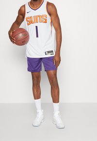 Persona con una camiseta de baloncesto de los Phoenix Suns y pantalones cortos morados sosteniendo un balón de baloncesto Spalding frente a un fondo blanco.