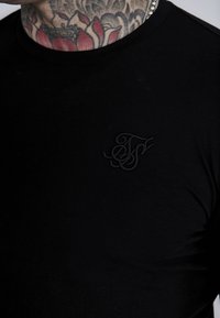 Crna majica s kratkim rukavima od glatkog materijala, s malim crnim izvezenim logotipom na prsima. Okrugli izrez, jednostavan dizajn, uski kroj.