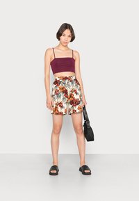 Top cropped em burgundy com alças finas, combinado com calções creme com um padrão floral tropical colorido. Sandálias de dedo pretas e uma bolsa pequena.