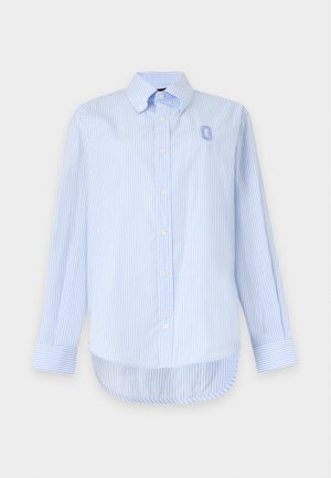 Personne debout, portant une chemise à rayures bleu clair, une mini-jupe en jean à motifs losanges patchwork, des ballerines noires, les mains dans les poches.