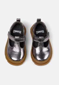 Lille børnesko i metallic sølv med Mary Jane-design, Velcro-stropper og tykke tan gummisåler, mærket "Camper" indeni.