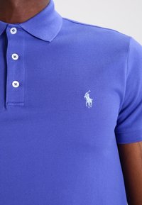 Paarse polo met een textuurstof, drie knoopsluiting en een lichtblauw geborduurd logo op de linkerborst.