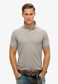 Lichtgrijze poloshirt met een korte kraag, een knoopsluiting met twee knopen, en een subtiel logo. Draag het met een geruit shirt om de taille, blauwe jeans en een horloge.
