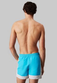 Costume da bagno da uomo, tessuto blu con bordo bianco. Vita elasticizzata e tasche laterali. Texture liscia, lunghezza fino a metà coscia. Vista posteriore.