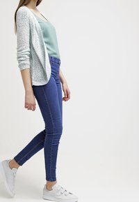 Lichtgroene tanktop, crèmekleurige gebreide cardigan, blauwe skinny jeans en witte sneakers. De cardigan heeft een open voorkant en een gestructureerde stof.