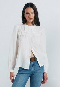 Weißes Langarm-Hemd mit Knopfleiste, verziert mit Spitzenbesatz und Faltendetails, kombiniert mit blauen hochgeschnittenen Jeans und einem braunen Gürtel.