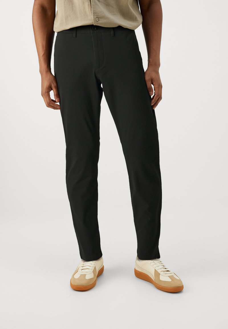 Black cotton trousers with a slim fit, flat front, and standard pockets. Paired with beige sneakers featuring textured accents.