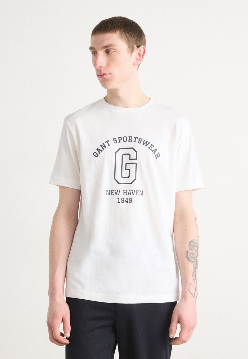 Valkoinen puuvillapaita, jossa on tummansinisellä painettuna "GANT SPORTSWEAR" ja "NEW HAVEN 1949". Lyhyet hihat, pyöreä kaula-aukko, rento istuvuus.
