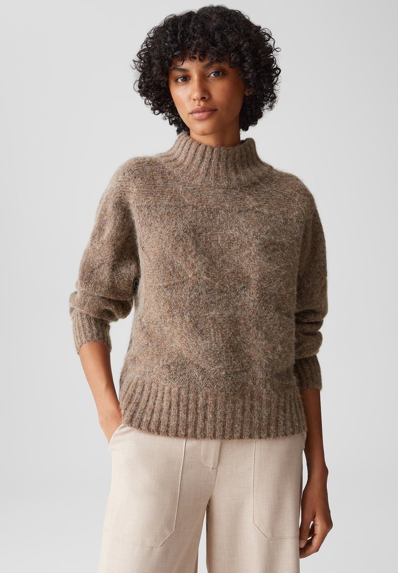 Maglione oversize marrone a maglia con colletto alto a coste, realizzato in un morbido tessuto testurizzato e con maniche a sbuffo. Indossato con pantaloni chiari.