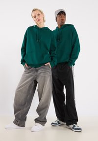 Grønn hettegenser, grå baggy jeans, svarte treningsbukser med striper, og sneakers i hvitt og grønt. Glatte teksturer, uformell stil.