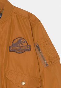Casaco bombário laranja feito de um tecido suave, com um emblema do Jurassic World no lado esquerdo do peito e um bolso com fecho de correr na manga.