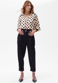Beige blouse met zwarte stippen, met knopen aan de voorkant en geraffineerde mouwen. Hoge taille zwarte broek met plooien en een opgerolde zoom. Zwarte hakken.