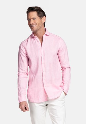 Man glimlachend, gekleed in een lichtroze overhemd met lange mouwen en witte broek, staand met één hand in de zak tegen een effen achtergrond.
