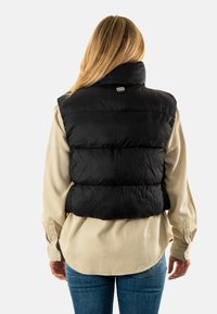 Chaleco acolchado negro con cuello alto, material ligero, diseño acolchado, llevado sobre una camisa de manga larga beige, con un logotipo de la marca en la parte posterior.