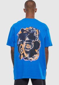 Blå bomulds T-shirt med et stort grafisk print på ryggen, der viser stiliserede figurer og ansigtstræk i sort og orange, omkranset af lys lilla.