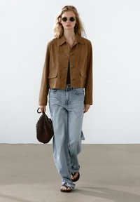Giacca cropped in suede marrone con due tasche frontali, abbinata a jeans blu chiaro e a una borsa in tessuto nera. Modella con occhiali da sole neri e sandali.
