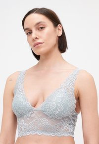 Krekls bralette gaiši zilā krāsā, ar ziedu rakstiem, dziļu V veida kakla izgriezumu un viļņainām malām. Mīksta tekstūra ar platām mežģīņu siksnām.