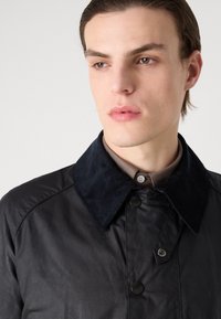 Veste noire avec un col en velours côtelé, dotée d'une fermeture à boutons à l'avant et d'une texture lisse ; portée par-dessus une chemise marron à col.
