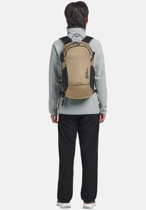 Persoon die met de rug naar de camera staat, draagt een lichtgrijs jack, zwarte broek, witte wandelschoenen en een beige rugzak met zwarte accenten en logo.