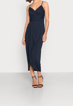 Kobieta ubrana w granatową sukienkę midi z kopertowym dekoltem na cienkich ramiączkach oraz czarne szpilki z paskiem wokół kostki, stojąca na tle jednolitego jasnego tła.