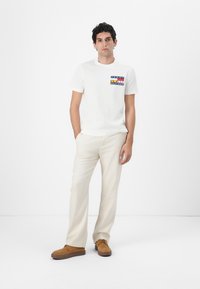 Witte t-shirt met korte mouwen en een gekleurde logoprint op de borst, gecombineerd met lichtbeige broek en bruine suède schoenen.