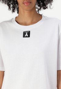 Baltas kokvilnas T-krekliņš ar apaļu kakla izgriezumu un īsām piedurknēm, ar melnu logotipa etiķeti centrā kakla rajonā. Mīksta tekstūra.