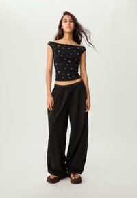 Mladá žena s dlouhými vlasy má na sobě černý crop top bez ramínek s kruhovými vzory, volné černé kalhoty a platformové sandály na bezvýznamném pozadí.