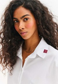 Biała koszula zapinana na guziki z kołnierzykiem, z małą bordową naszywką z logo. Gładka tkanina i luźny krój, podkreślające neutralne odcienie.