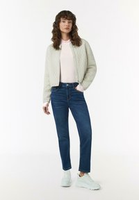 Hellgraue Kabelstrick-Bomberjacke, pinkes Langarmoberteil, blaue gerade Jeans und weiße Sneakers, stehend vor einem einfarbigen Hintergrund.