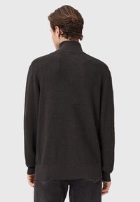 Man in een zwarte ribgebreide trui met hoge kraag, weergegeven van achteren tegen een effen lichtgrijze achtergrond.
