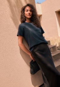 Donkerblauwe top met korte mouwen gecombineerd met een strakke zwarte rok. Het model houdt een zwarte clutch met textuur vast, tegen een neutrale achtergrond.