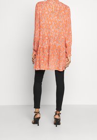 Personne portant un tunique florale orange à manches longues, un pantalon skinny noir et des sandales à talons hauts noires, se tenant devant un fond blanc.