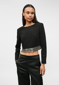 Μαύρη κοντή μπλούζα με μακριά μανίκια από λείο ύφασμα, με μεταλλική αλυσίδα στο τελείωμα, συνδυασμένη με κομψά μαύρα παντελόνια.