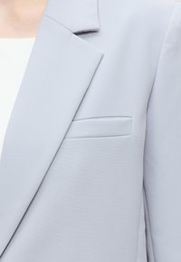 Détail du revers de blazer gris clair et de la poche poitrine sur une personne portant un haut blanc dessous.