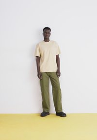 Beige pamut póló zsebbel, olíva zöld laza szabású nadrággal és fekete slip-on cipővel egy sárga padlón.