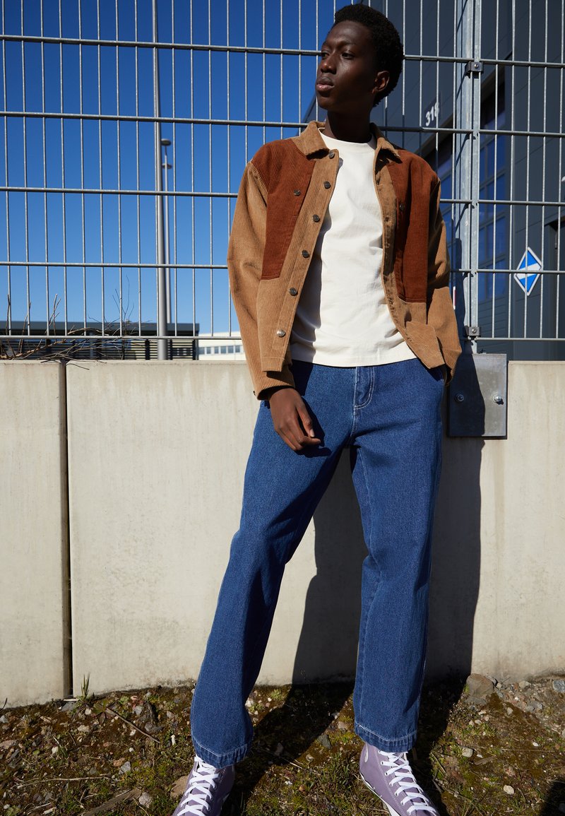 Brown and tan corduroy shirt jacket over a white T-shirt, paired with blue straight-leg jeans and purple high-top sneakers.