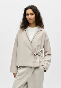 Femme aux cheveux bouclés portant une veste portée beige clair nouée à la taille sur une chemise blanche, associée à un pantalon large assorti.