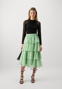 Top negru cu mânecă lungă, fustă verde mentă stratificată cu volane și sandale negre cu bretele. Modelul ține o geantă neagră.