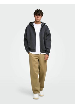Jeune homme debout portant un sweat à capuche zippé gris foncé, un t-shirt blanc, un pantalon beige à jambes larges et des baskets blanches sur un fond gris clair.