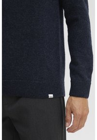 Pull en laine bleu marine avec des poignets et un ourlet côtelés, arborant une petite étiquette de marque blanche. La manche est présentée à côté d'un pantalon gris.