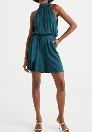 Femme portant une combinaison short sans manches couleur sarcelle avec ceinture à la taille et sandales blanches à talons hauts, debout devant un fond uni.
