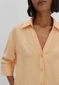 Camicia button-up arancione chiaro in tessuto texture, con scollatura a V e maniche a tre quarti, accentuata da un colletto semplice.