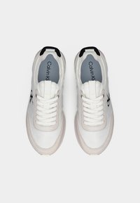 Ζευγάρι λευκά αθλητικά παπούτσια Calvin Klein με κορδόνια μπροστά και μαύρες λεπτομέρειες, σε οπτική από πάνω πάνω σε απλό φόντο.