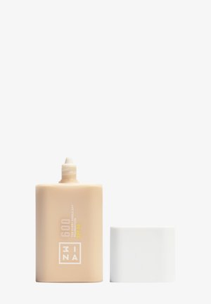 Foundationfles in lichtbeige plastic, met een witte dop. Het etiket bevat tekst in geel en wit. De bovenkant heeft een nozzle voor het doseren.