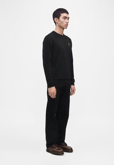 Jeune homme debout de profil portant une chemise noire à manches longues, un pantalon noir et des chaussures marron sur un fond clair uni.