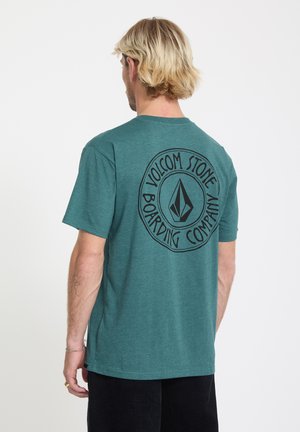 Homme blond portant un t-shirt turquoise avec un grand logo circulaire noir « Volcom Stone Boarding Company » dans le dos, debout sur un fond blanc.