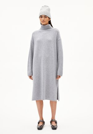 Hellgraues Strickkleid mit hohem Kragen, langen Ärmeln, seitlichen Schlitzen und losem Schnitt. Kombiniert mit einer grauen gerippten Mütze und schwarzen Schuhen.