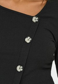 Le haut noir à côtes présente un décolleté asymétrique et trois grands boutons marbrés circulaires sur le côté. Texture douce évidente.