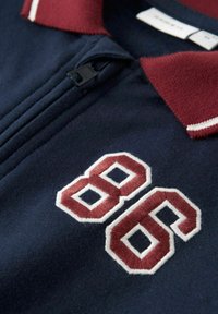 Tumesinine polo-särk marooni krae ja valgete ääristega, mille esipoolt kaunistab marooni ja valgega bordüüritud "88", ning millel on lukuga sulgemine.